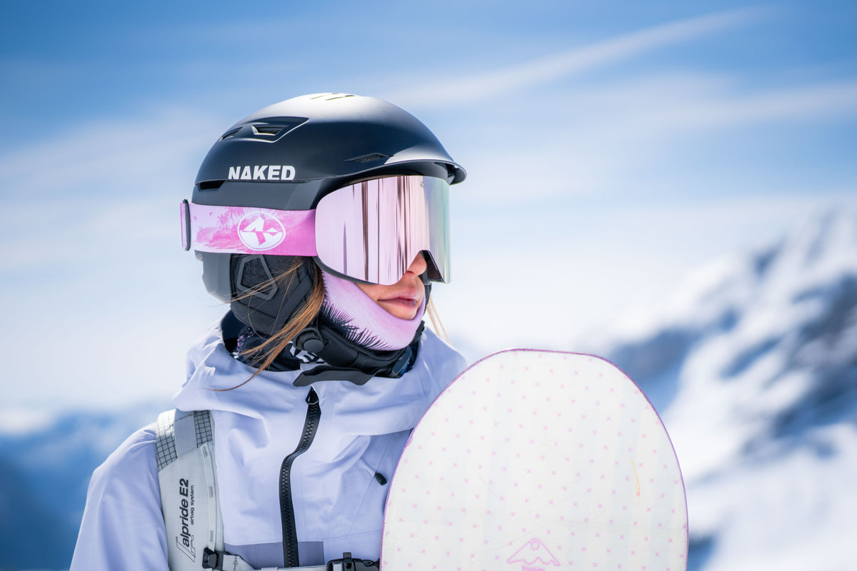 Eine Frau trägt einen schwarzen Skihelm und eine rosa Skibrille, sie hält ein Snowboard in der Hand