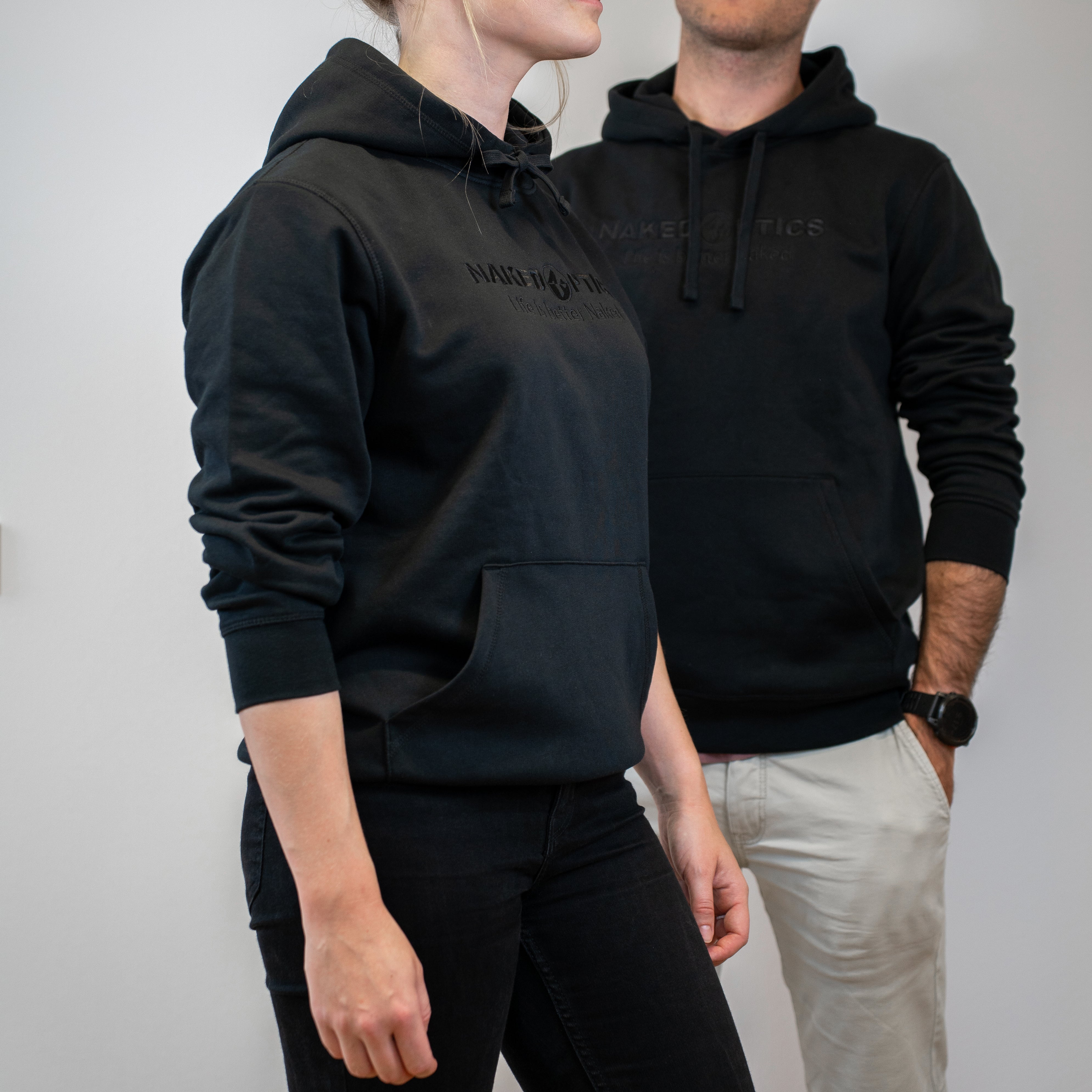 Zwei Personen, die NAKED Optics Unisex Hoodie in schwarz mit gesticktem Logo tragen, stehen nebeneinander vor einem schlichten hellen Hintergrund.
