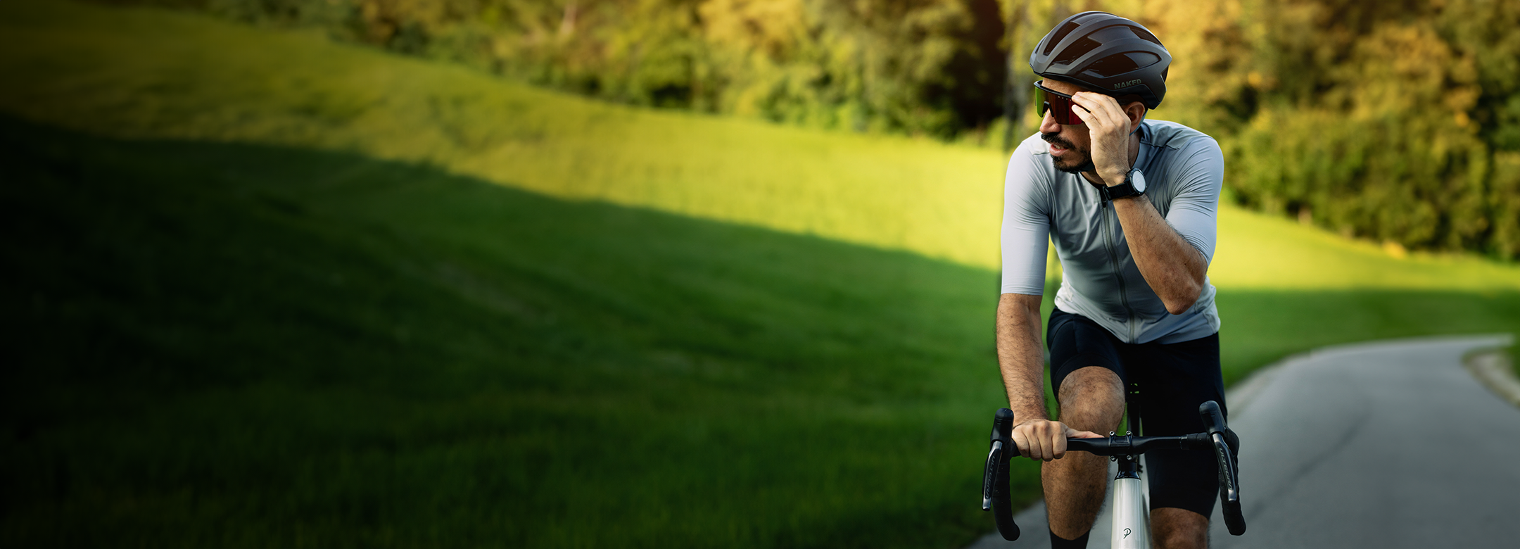 Ein Mann fährt mit dem Fahrrad auf einer Straße, er trägt einen Helm und eine Sonnenbrille; im Hintergrund sind grünes Gras und Bäume zu sehen.