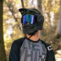 Una persona con un casco da bicicletta nero e occhiali protettivi, che indossa una maglia grigia e sfoggia gli occhiali da MTB di alta qualità “The ZEND” di NAKED Optics, si trova in un bosco illuminato dal sole.