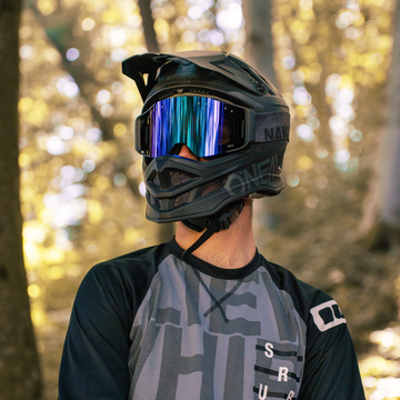 Eine Person mit Vollvisierhelm und NAKED Optics MTB Brille mit magnetischen Wechselgläsern steht in einem sonnenbeschienenen Wald, bekleidet mit einem grau-schwarzen Trikot.