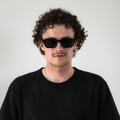 Ein Mann mit lockigen Haaren trägt die schwarze Sonnenbrille “The IRIS” von NAKED Optics mit polarisierten Gläsern und UV400-Schutz sowie ein schwarzes Shirt und steht vor einem schlichten weißen Hintergrund.