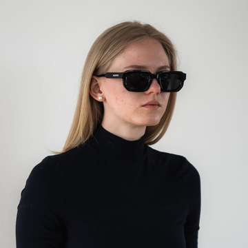 Frau mit glattem blondem Haar trägt eine NAKED Optics Sonnenbrille mit polarisierten Gläsern und UV400-Schutz sowie einen schwarzen Rollkragenpullover vor schlichtem Hintergrund.