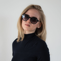A woman with long blonde hair, wearing “The IZZY” UV400 sunglasses by NAKED Optics featuring an acetate frame, and a black turtleneck, poses against a plain background.