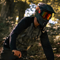 A person wearing a helmet and NAKED Optics The LINE MTB Brille rides a bike through a sunlit forest.