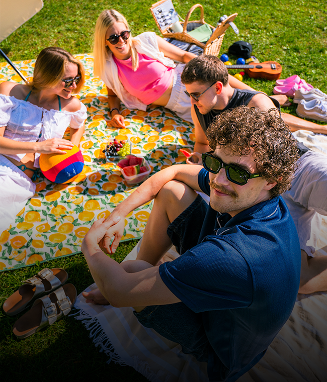 Vier Freunde entspannen sich draußen auf einer Picknickdecke mit Snacks, Sonnenbrillen und bei sonnigem Wetter.