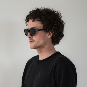 Mann mit lockigem Haar trägt eine NAKED Optics LUNA Sonnenbrille mit polarisierten Gläsern und ein schwarzes Shirt, stehend vor weißem Hintergrund.