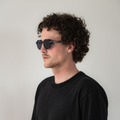 A man with curly hair, dressed in a black shirt, stands against a plain white background wearing The LUNA by NAKED Optics, effortlessly showcasing the stylish appeal of this lifestyle eyewear featuring polarized lenses.