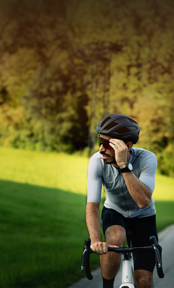 Ein Mann mit Helm fährt auf einem Weg Fahrrad, richtet seine Sonnenbrille zurecht, im Hintergrund sind grünes Gras und Bäume zu sehen.