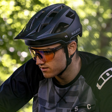 Ein Radfahrer, der eine NAKED Optics Sonnenbrille und den RIDGE MTB Helm trägt, fährt im Freien mit grünem Laub im Hintergrund.