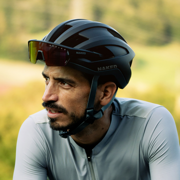 Ein Mann mit einem schwarzen NAKED Optics Rennrad-Helm und einer Sonnenbrille trägt ein graues Trikot im Freien mit einer Grünfläche hinter ihm. Die aerodynamische Form des Helms steigert sowohl seinen Stil als auch seine Leistung.