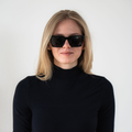 A woman with long blonde hair, dressed in a black turtleneck, is wearing The MUSE sunglasses by NAKED Optics—a stylish unisex model with UV400 protection and polarized lenses—while standing against a white wall.