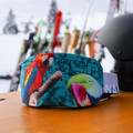 A colorful ski mask with parrot images rests on a wooden table; nearby, a vibrant NAKED Optics Skibrillen Schutz (Überzieher) adds flair to the ski gear, while snow glistens in the background.
