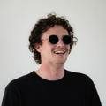 A man with curly hair and sunglasses smiles, wearing a black shirt against a plain white background. He sports the stylish NAKED Optics The PARKER Metallrahmen Sonnenbrille, which offers reliable UV400 Schutz.