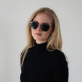 A woman with long blonde hair wears a stylish black Metallrahmen-Sonnenbrille—the PARKER by NAKED Optics—with a black turtleneck against a plain background. The PARKER Sonnenbrille offers UV400 Schutz for ultimate eye protection.