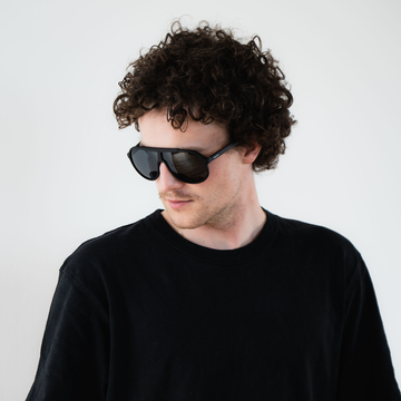 Mann mit lockigem Haar, der eine NAKED Optics PILOT Sonnenbrille mit UV400-Beschichtung und ein schwarzes Hemd trägt, blickt nach links.