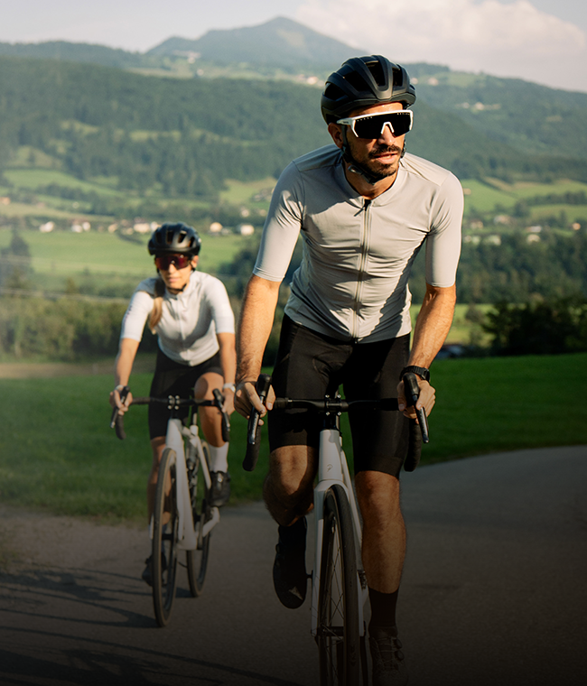 Zwei Radfahrer fahren auf einer Landstraße, im Hintergrund sind grüne Hügel und Berge zu sehen; sie tragen Helme und Sonnenbrillen.
