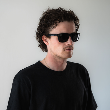 Ein Mann mit lockigem Haar, der ein schwarzes T-Shirt und eine NAKED Optics Sonnenbrille mit polarisierten Gläsern trägt, steht vor einem schlichten hellen Hintergrund.