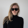 Eine Frau mit langen blonden Haaren, die die Sonnenbrille „The SLUSH“ von NAKED Optics mit polarisierten Gläsern trägt und einen schwarzen Rollkragenpullover, steht vor einer weißen Wand – perfekt gestylt für Outdoor-Aktivitäten.