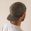A man is shown from behind against a beige background, wearing a gray NAKED Optics Cap Snapback featuring 3D Stick embroidery on the back.