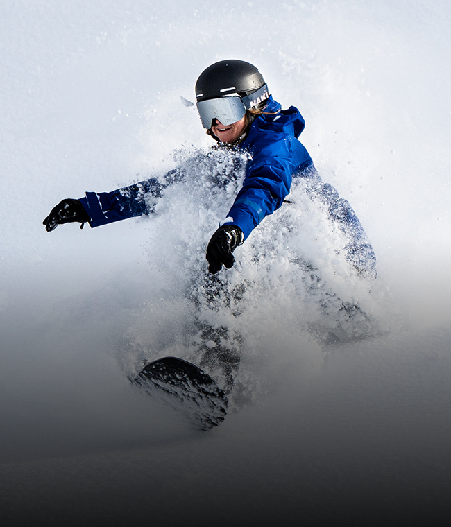 Eine Person fährt mit hoher Geschwindigkeit auf einem Snowboard durch den Pulverschnee; sie trägt eine blaue Jacke, einen schwarzen Helm und eine Skibrille. Dabei spritzt Schnee hoch.