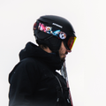 Eine Person in Skikleidung mit einer großen Skibrille, dem Modell „The NOVA“ von NAKED Optics, und einem Bärenaufkleber auf dem Helm steht vor einem weißen Hintergrund und blickt nach rechts. Die Skibrille ist mit einer praktischen Anti-Beschlag-Beschichtung ausgestattet.