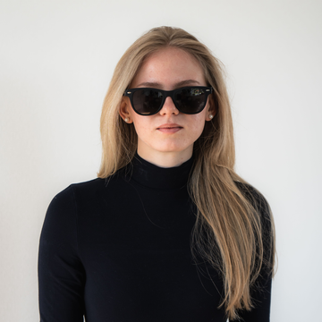 Frau mit langen blonden Haaren trägt NAKED Optics schwarze Sonnenbrille mit UV-Schutz und einen schwarzen Rollkragenpullover, stehend vor einem weißen Hintergrund.