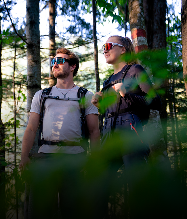 Zwei Menschen mit Rucksäcken und Sonnenbrillen stehen in einem sonnendurchfluteten Wald, umgeben von grünen Blättern und Bäumen.