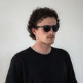 A man with curly hair, dressed in a black shirt and wearing black sunglasses, stands against a plain white background, modeling The WHOOP polarized sunglasses with UV400 protection from NAKED Optics.