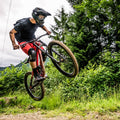 A mountain biker in red shorts launches off a dirt trail amidst lush green trees and foliage, confidently wearing NAKED Optics The RIFT goggles for crystal-clear vision on the ride.