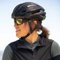 A smiling person wearing a bike helmet, a black vest, and The CHARGE Fullframe Sportsonnenbrille with UV400 Schutz from NAKED Optics looks to the left outdoors.