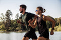In una giornata di sole, due persone fanno jogging in riva al lago; entrambe indossano gli eleganti occhiali da sole sportivi NAKED Optics The RUSH Halfframe con protezione UV400, che completano i loro outfit sportivi. Sullo sfondo si vedono degli alberi.
