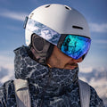 Un uomo in tenuta da sci con un casco bianco e gli occhiali da neve NAKED Optics The STORM blu, di tipo sferico e con rivestimento antiappannamento, con le montagne innevate che si riflettono nelle lenti, in una scena all’aperto.