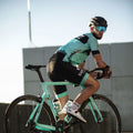 A cyclist in teal and black gear rides a teal road bike, wearing a helmet and NAKED Optics’ The FALCON Sportsonnenbrille with UV400 Schutz.