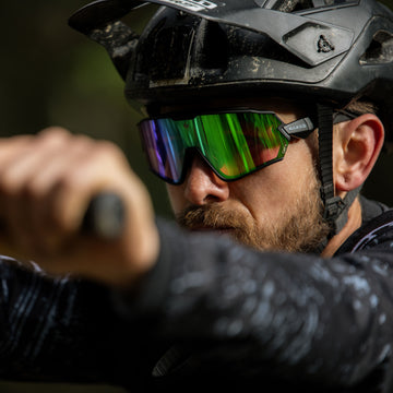 Ein bärtiger Radfahrer mit Helm und reflektierendem NAKED Optics Sportbrille umklammert den Lenker und konzentriert sich auf die Strecke vor ihm.