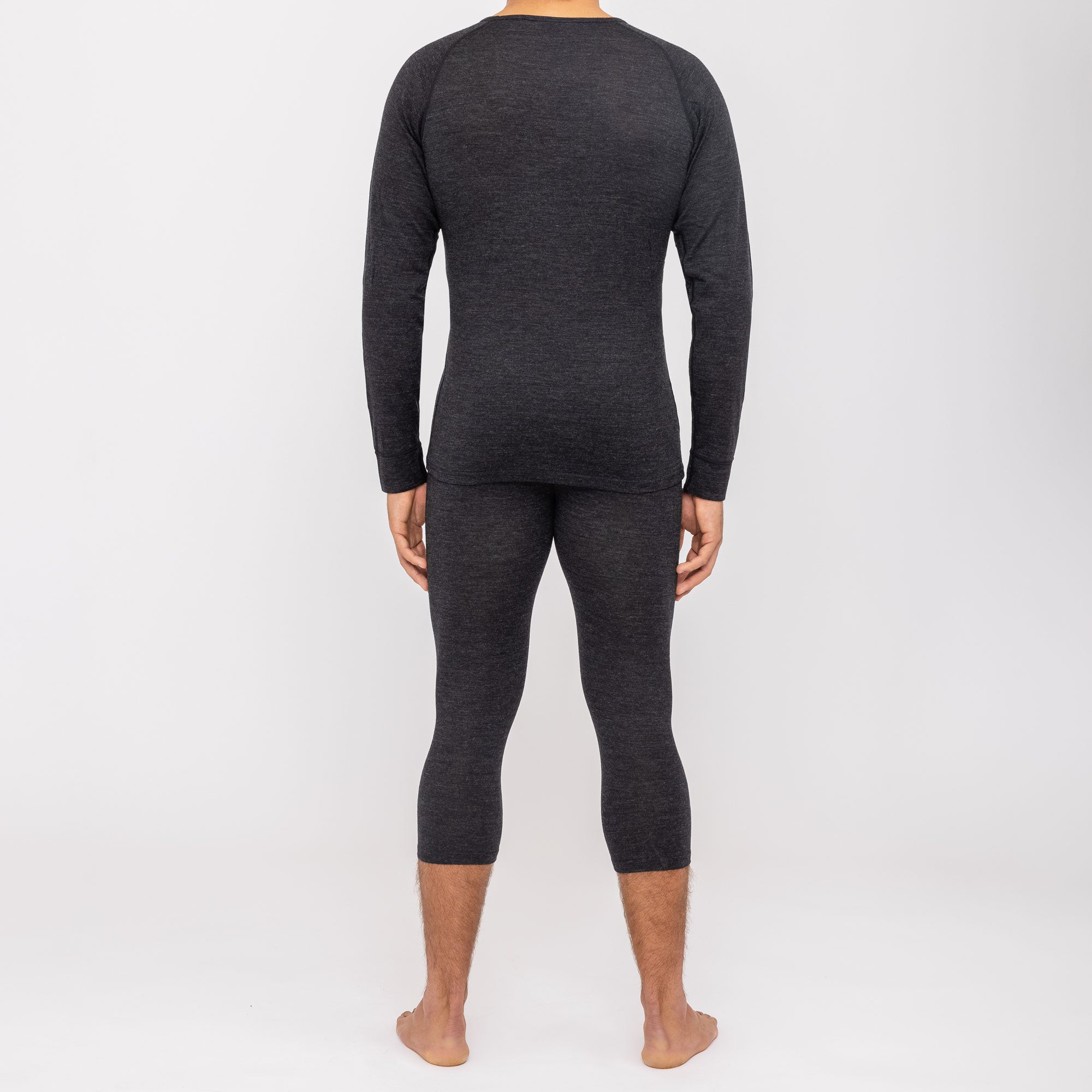 Eine Person steht barfuß, mit dem Gesicht nach außen, in dunkler langärmeliger NAKED Optics Merino Unterwäsche und passenden Leggings auf weißem Grund.