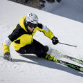 Ein Skifahrer in gelber Jacke und mit Helm fährt auf einem verschneiten Hang eine scharfe Kurve, wobei der Schnee hochspritzt. Er trägt dabei die Skibrille „The NOVA“ von NAKED Optics mit Antibeschlagbeschichtung für klare Sicht.