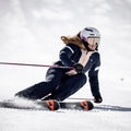 Ein Skifahrer in marineblauer Ausrüstung und mit einem weißen Helm, der die NOVA-Skibrille mit Anti-Fog-Beschichtung von NAKED Optics trägt, fährt auf einem verschneiten Hang bergab und macht dabei scharfe Schwünge.