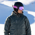 Ein:e Skifahrer:in mit Helm und der sphärischen Skibrille The STORM von NAKED Optics mit Anti-Fog-Beschichtung steht auf verschneiten Hängen und blickt nach links.