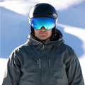 Person in ski gear, helmet, and reflective goggles—The STORM by NAKED Optics with sphärischer Skibrille and Anti-Fog-Beschichtung—standing in a snowy outdoor setting under sunlight.