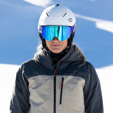 Eine Person in Skikleidung und mit Helm trägt eine reflektierende, rahmenlose Kinder-Skibrille von NAKED Optics mit Anti-Beschlag-Beschichtung und steht auf einer sonnigen, verschneiten Piste.