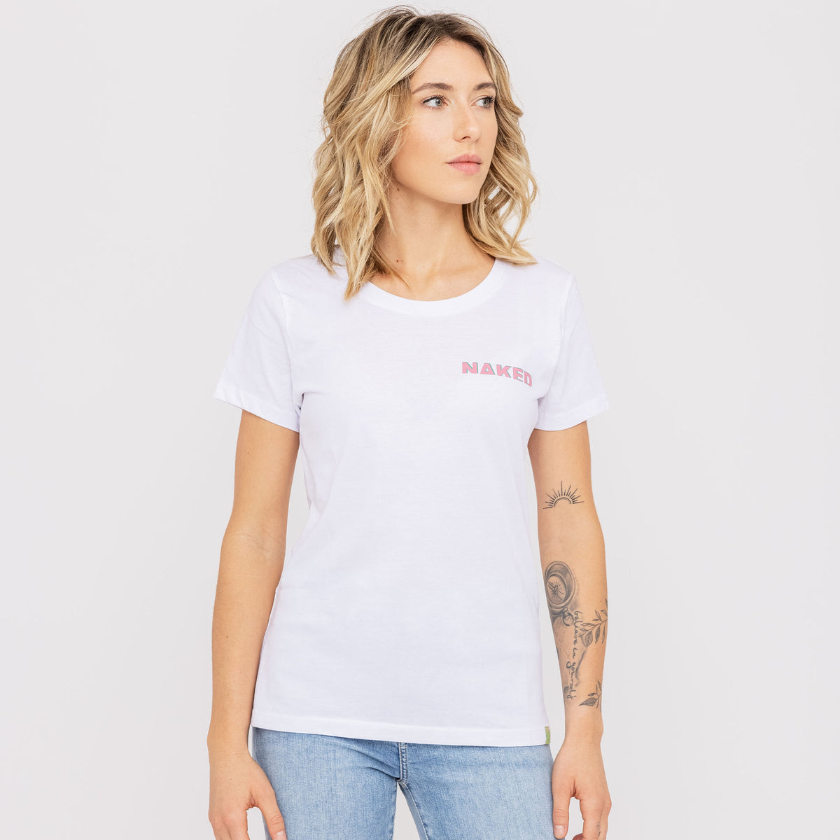 Une femme se tient debout devant un fond uni, vêtue d'un t-shirt NAKED Optics au logo bicolore, confectionné en coton bio tout doux, qu'elle porte avec un jean bleu clair.