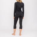 A woman stands barefoot, facing away, on a white background, wearing a black long-sleeve top and cropped leggings. This stylish set is the Thermo-Unterwäsche Damen (100% Merino) by NAKED Optics, expertly crafted from soft Merino Wolle for exceptional comfort and breathability as premium Funktionswäsche Damen.