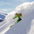 Uno sciatore in tenuta verde effettua una curva brusca, sollevando neve fresca su un pendio soleggiato, mentre indossa gli occhiali TROOP EVO di NAKED Optics, dotati di rivestimento antiappannamento per una visione nitida.