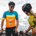 Two cyclists in helmets and jerseys smile and chat while holding their bikes, both sporting the stylish “The FALCON” Sportsonnenbrille with UV400 Schutz from NAKED Optics, with majestic mountains rising in the background.