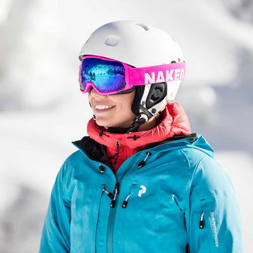 Lächelnder Skifahrer in blauer Jacke, weißem Helm und rosafarbener NAKED Optics-Brille mit verspiegelten Gläsern (100 % UV-Schutz) vor einer verschneiten Kulisse.