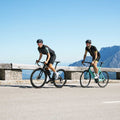 Zwei Radfahrer fahren unter strahlend blauem Himmel auf einer asphaltierten Bergstraße; beide tragen die RUSH Halfframe von NAKED Optics mit UV400-Schutz für optimalen Augenschutz, im Hintergrund sind Berge zu sehen.